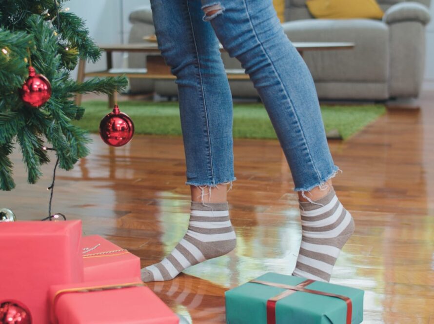 Foto desde un suelo de madera. Se aprecia el pie de un árbol de navidad decorado con bolas y luces, debajo de él unos regalos y al lado de estos unos pies en puntillas de una mujer, se puede intuir que está colocando algún adorno en el árbol.