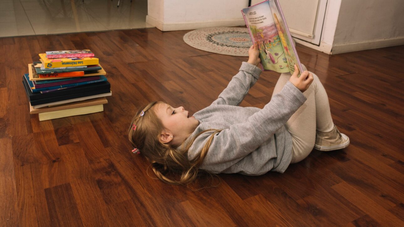 Niña leyendo un cuento infantil, tumbada en el suelo de madera de lo que parece un salón