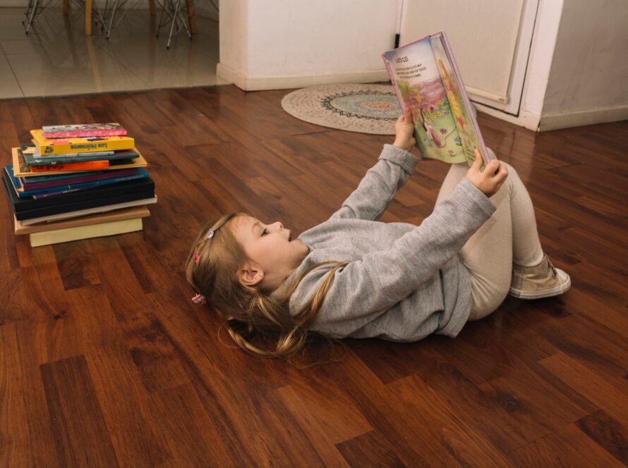 Niña leyendo un cuento infantil, tumbada en el suelo de madera de lo que parece un salón
