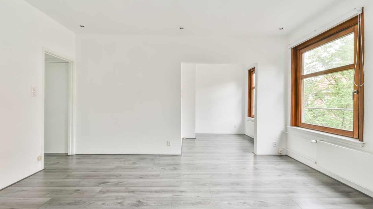 Interior de una habitación con paredes blancas, ventanas al exterior y tarima de color gris en el suelo.